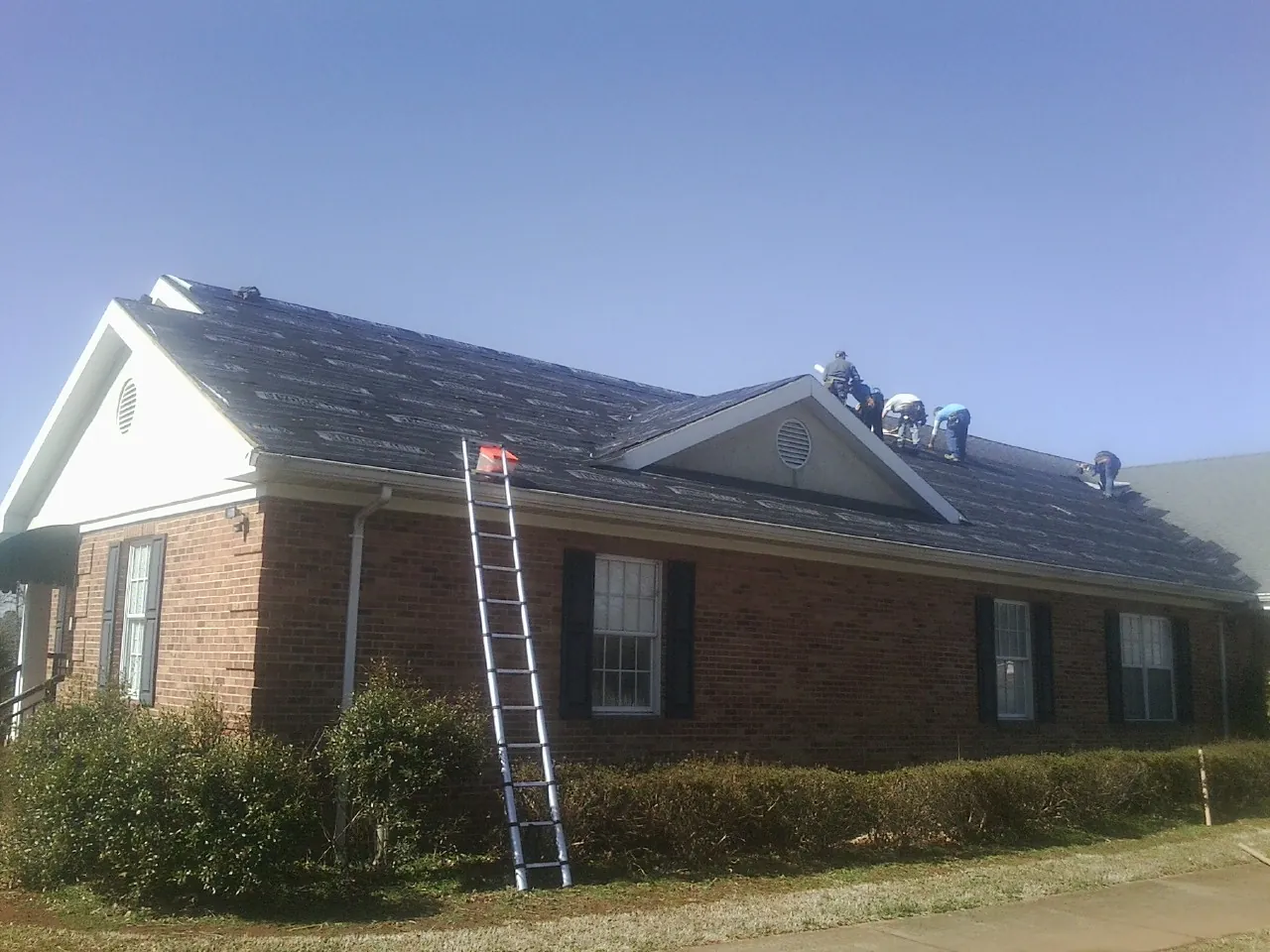 Crew tearing shingles off a brick ranch roof with a ladder set — Metal Roof Repair work by our crew in Long Pine