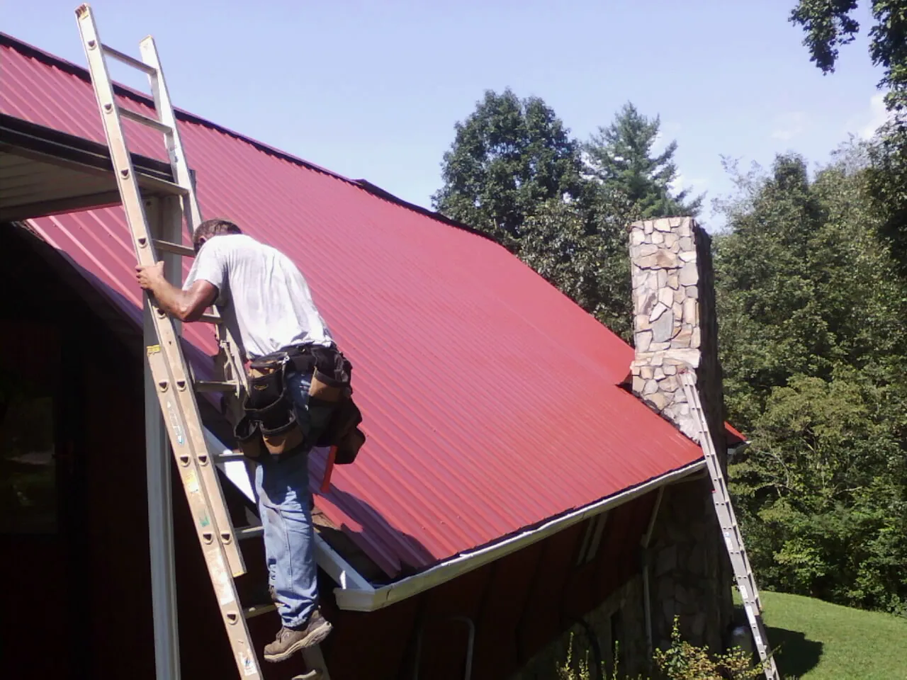 Roofer on a ladder beside a red metal roof and stone chimney — Soffit & Fascia Repair by Roofing Brothers in Long Pine